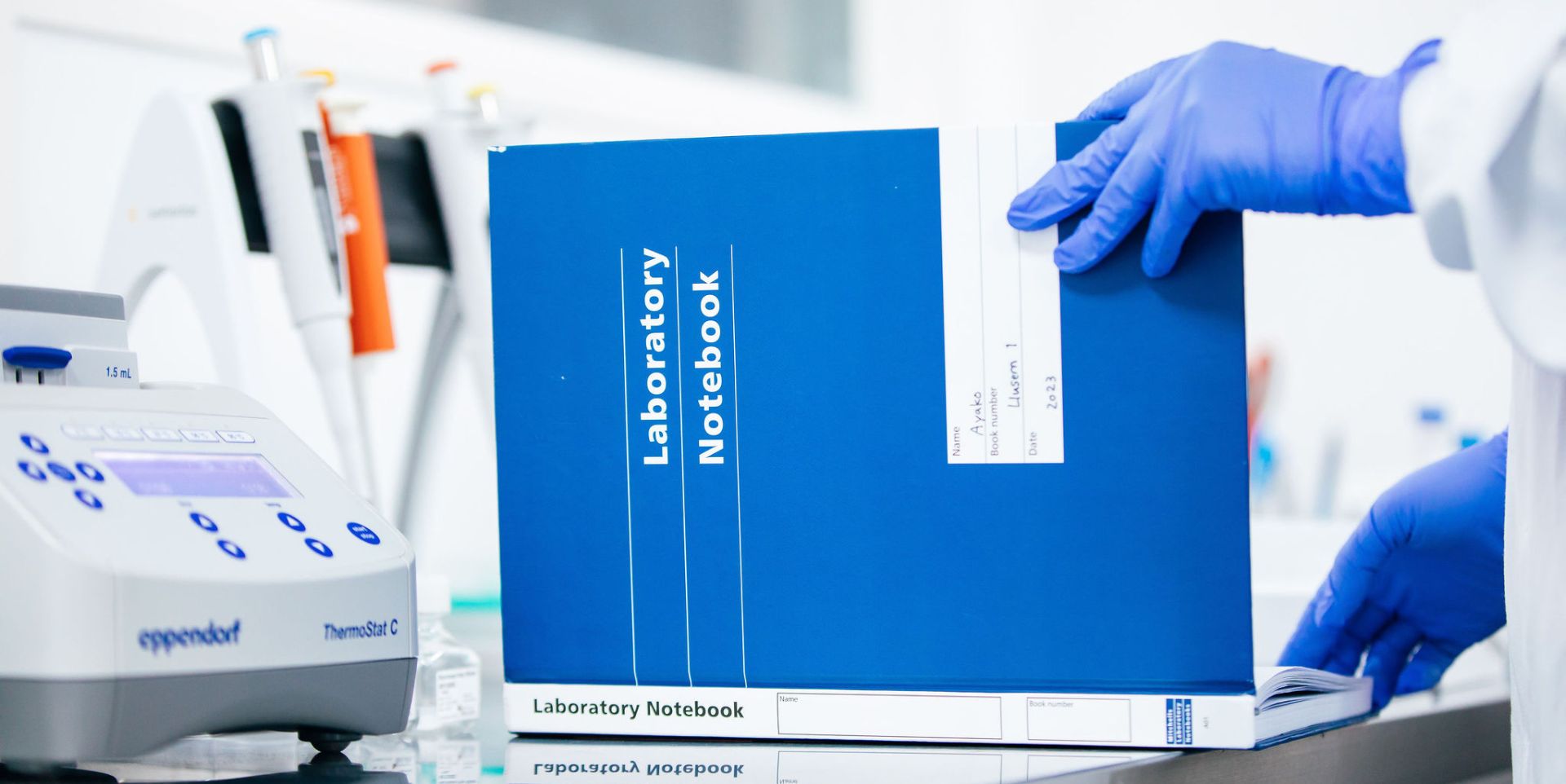 Moving into a new lab space: image depicts a scientist in a lab coat with a blue glove opening a bright blue notebook titled "Laboratory notes". Lab equipment can be seen to the left in the background.