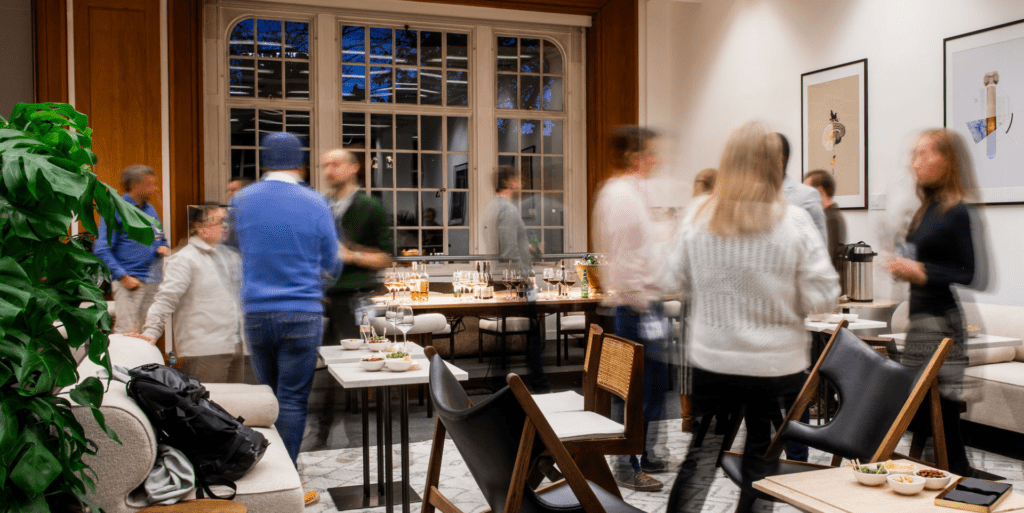 Life sciences investment trends: images depicts a room of people at an evening event. A green plant is in focus in the foreground, and a large window is in focus in the background. There in a motion blur effect on the people to show movement.