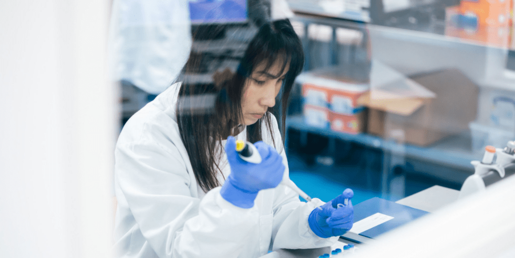 Government initiatives for life sciences: image depicts a scientist working in a lab. She has long black hair and is wearing a white lab coat and blue gloves. She is holding a pipette in her hands, and has a look of concentration on her face.